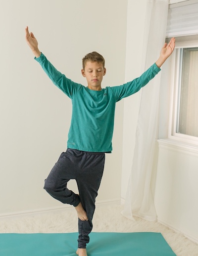 A grade-schol boy in a green sweatshirt doing a yoga pose with arms in the air